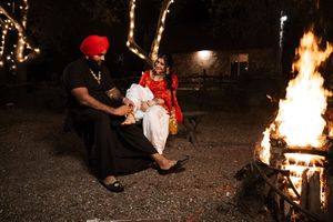 Two people sit together near a glowing campfire at night, with one wearing traditional red attire.