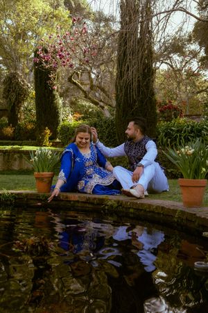 Couple in traditional Indian attire sitting by a garden pond, woman in blue salwar kameez, man in white kurta.