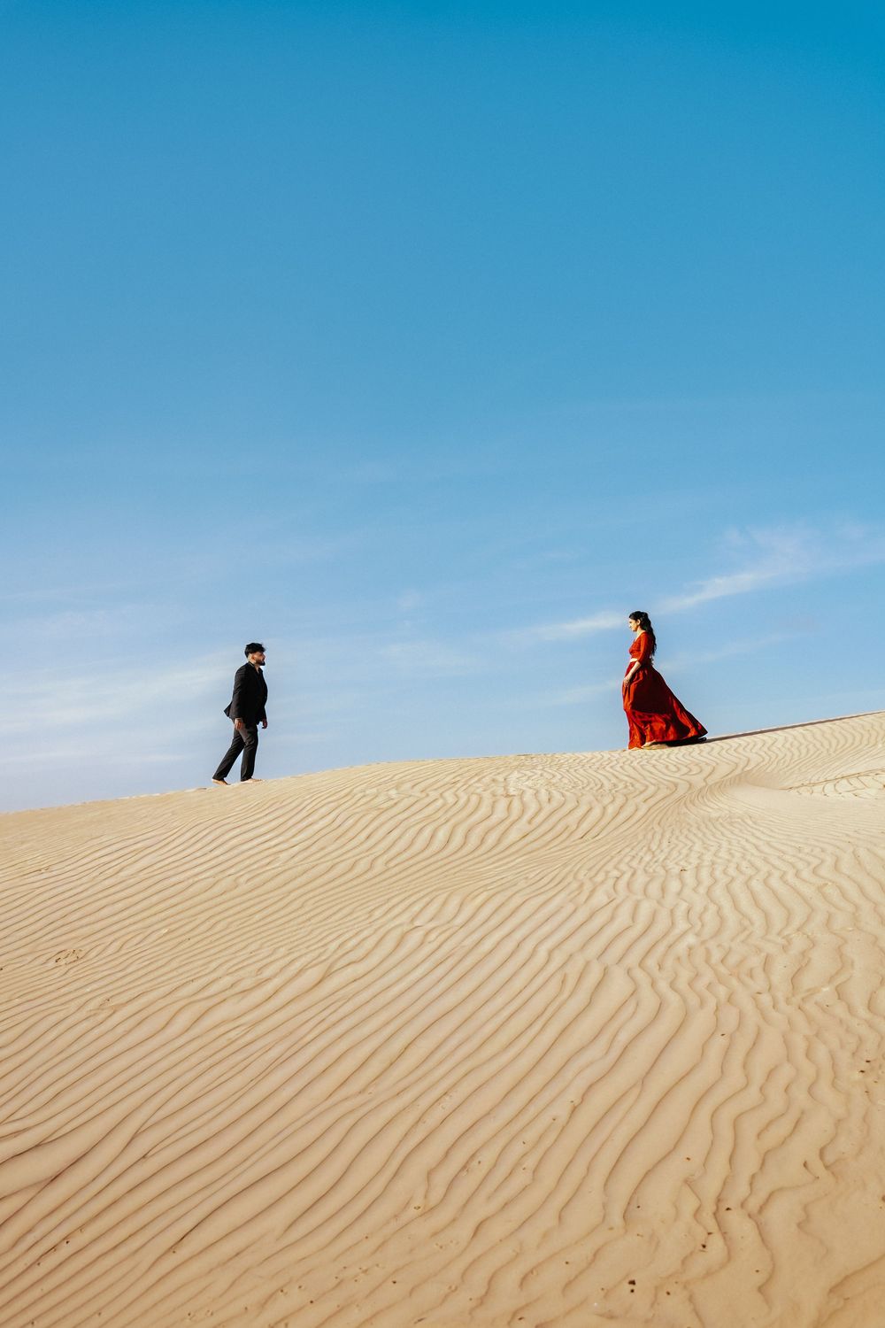 A couple in formal attire walks along a vast sandy desert dune under a clear blue sky.