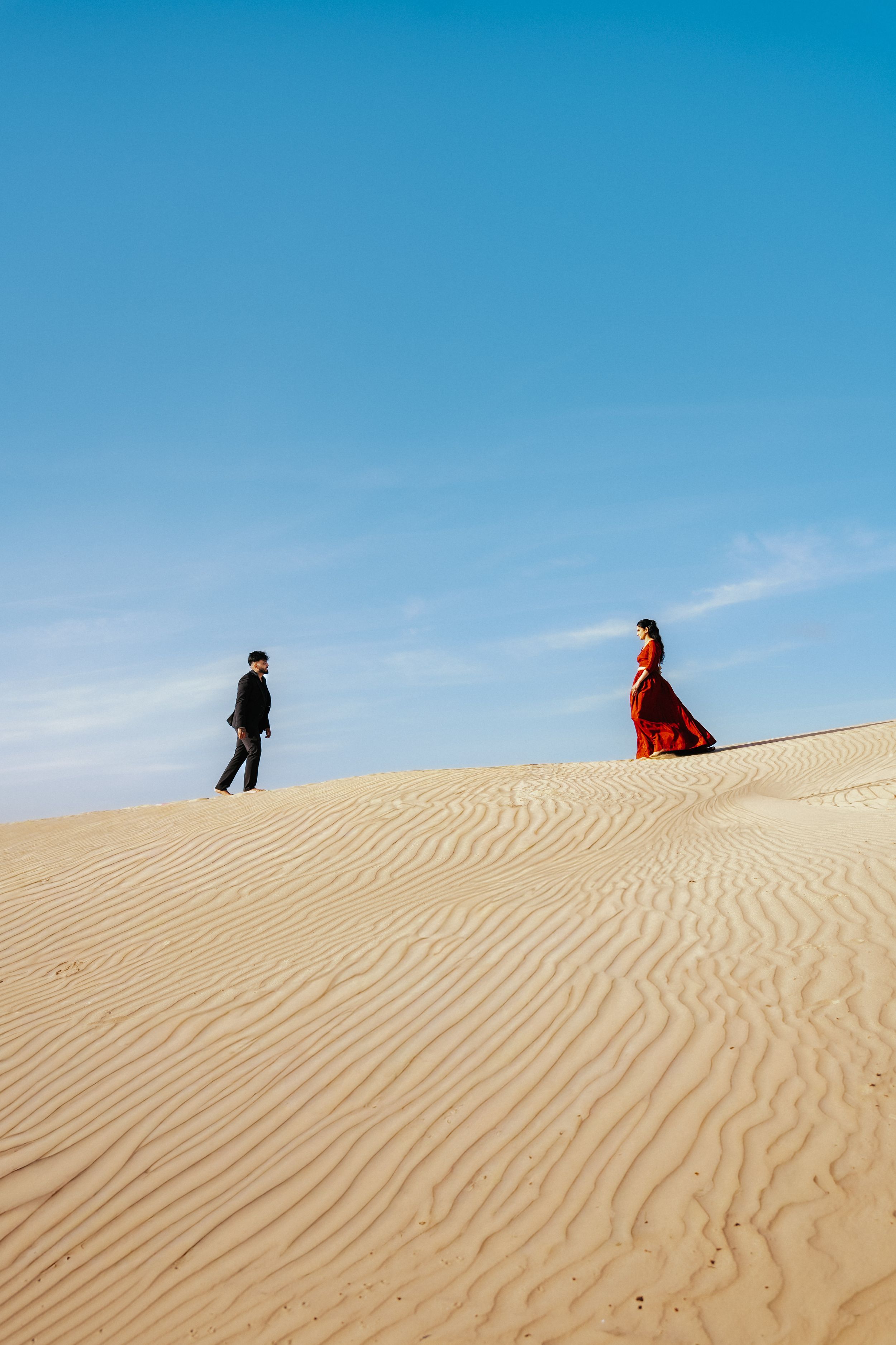 A couple in formal attire walks along a vast sandy desert dune under a clear blue sky.