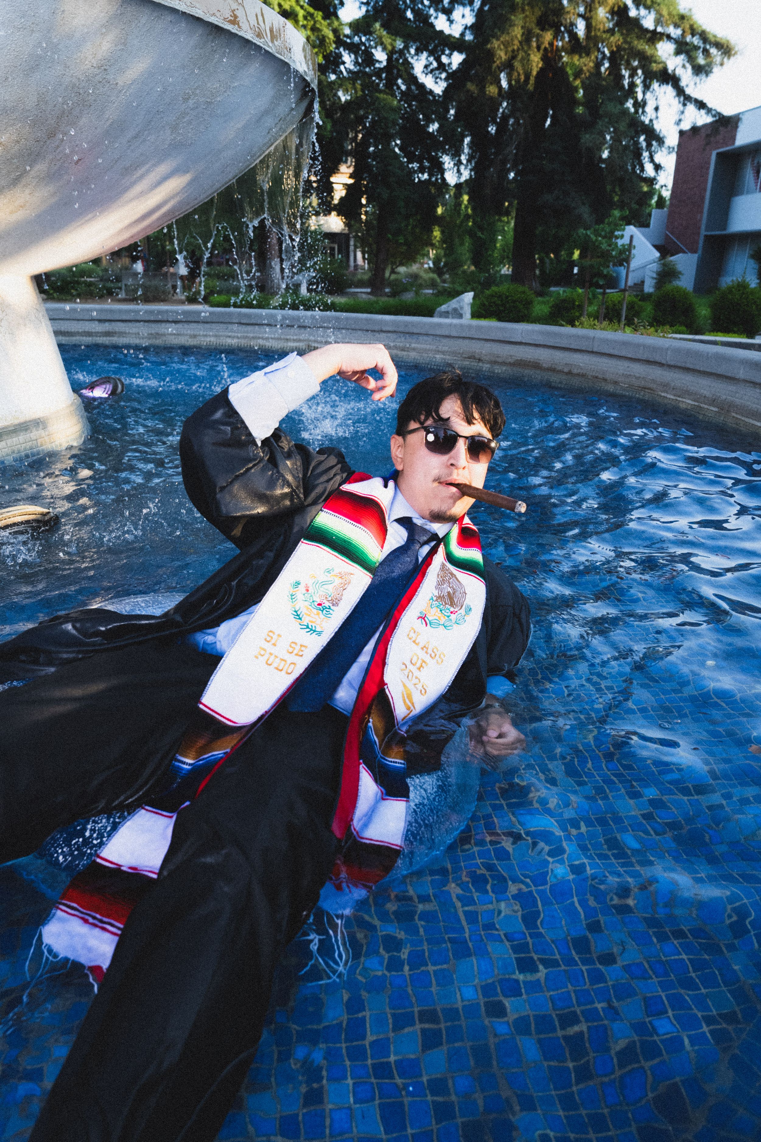 A person wearing sunglasses and a suit floats in a blue tiled fountain with spraying water.