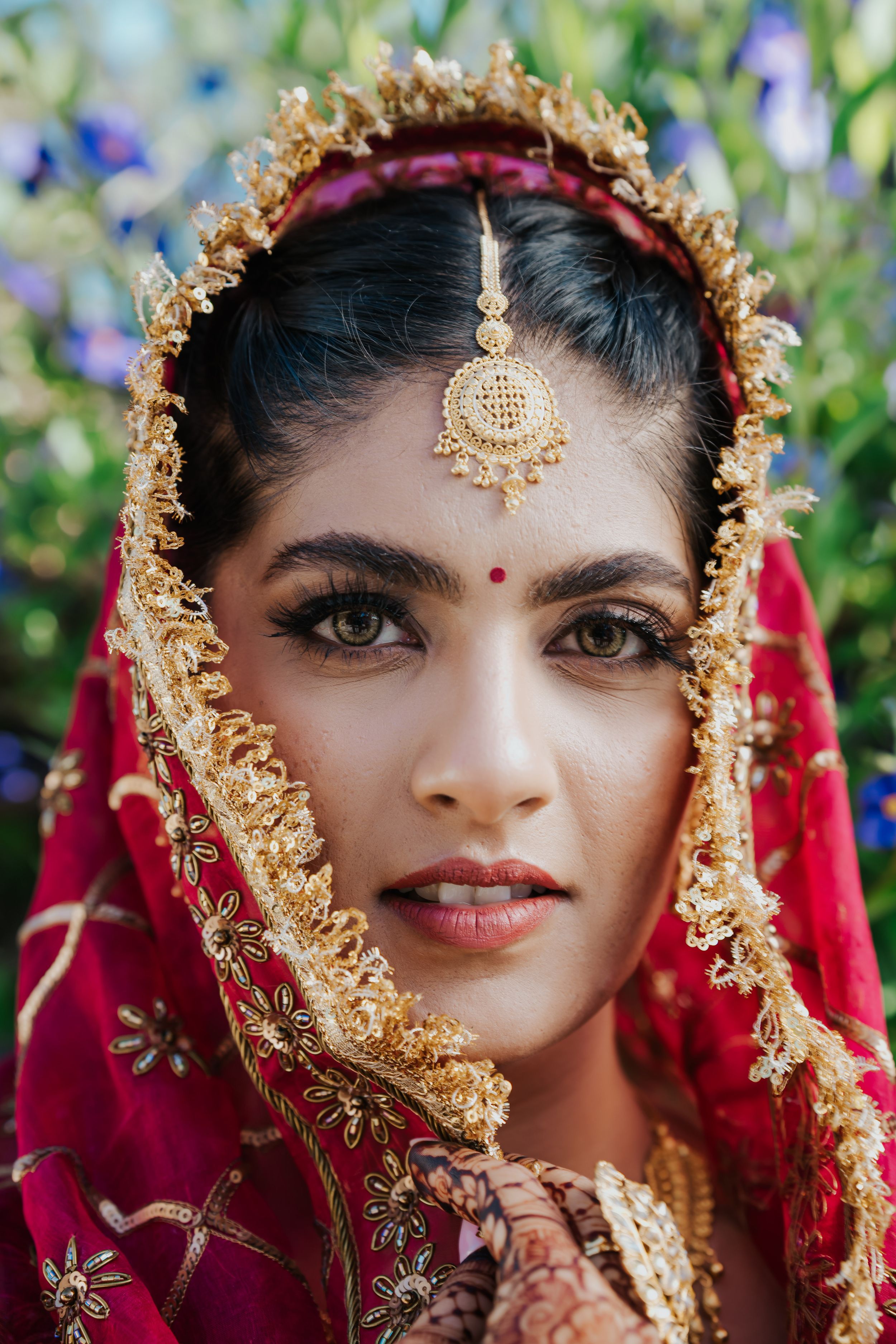 Traditional Indian bride wearing a red and gold embroidered dupatta with tikka and ornate jewelry against floral background.