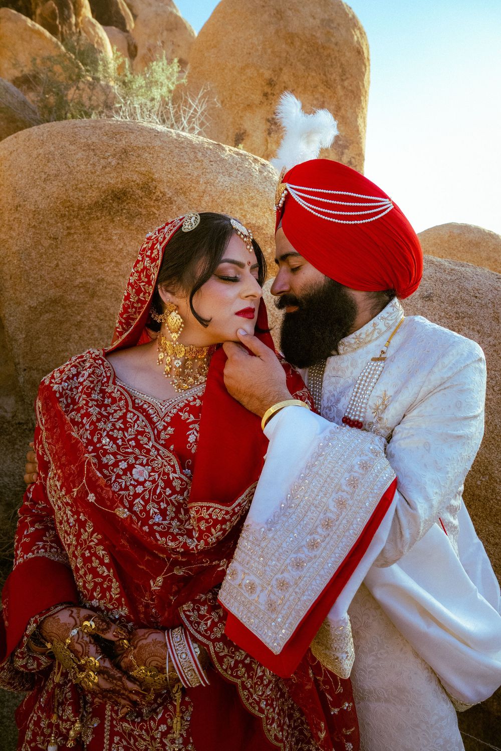 Sikh bride in red lehenga and groom in white sherwani with red turban share a romantic moment outdoors.