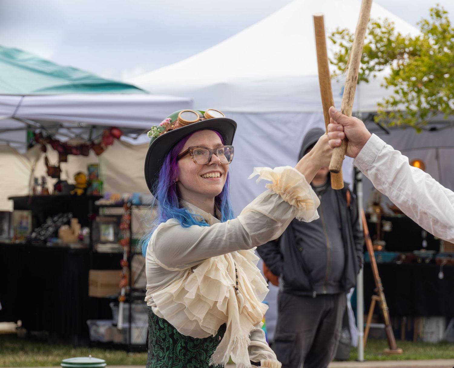 A young woman with purple and blue hair and steampunk costume clashes sticks with a dance partner. There are market stall in the background.