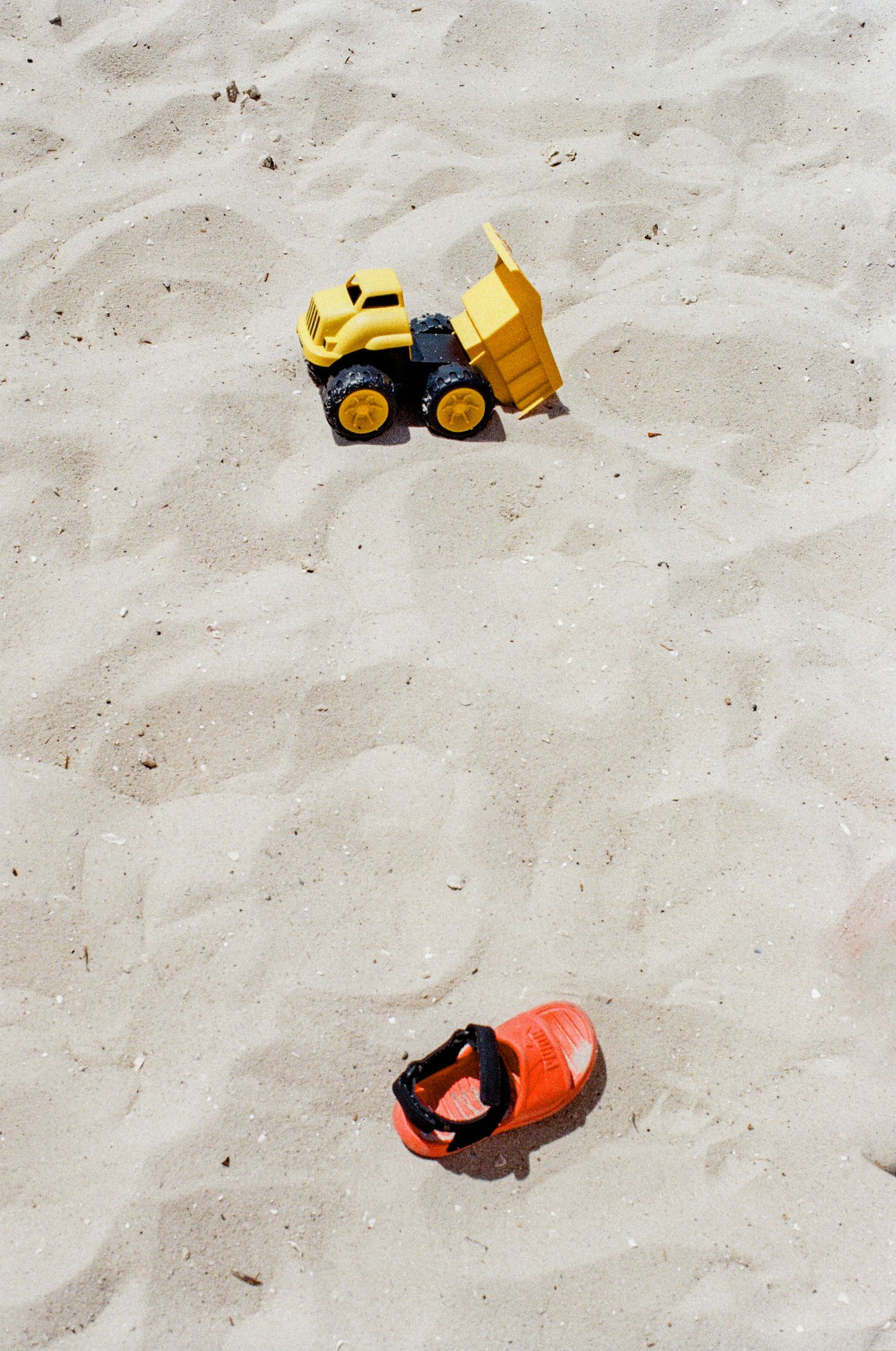 A yellow toy bulldozer and orange sandal sit isolated on smooth beige sand at the beach.