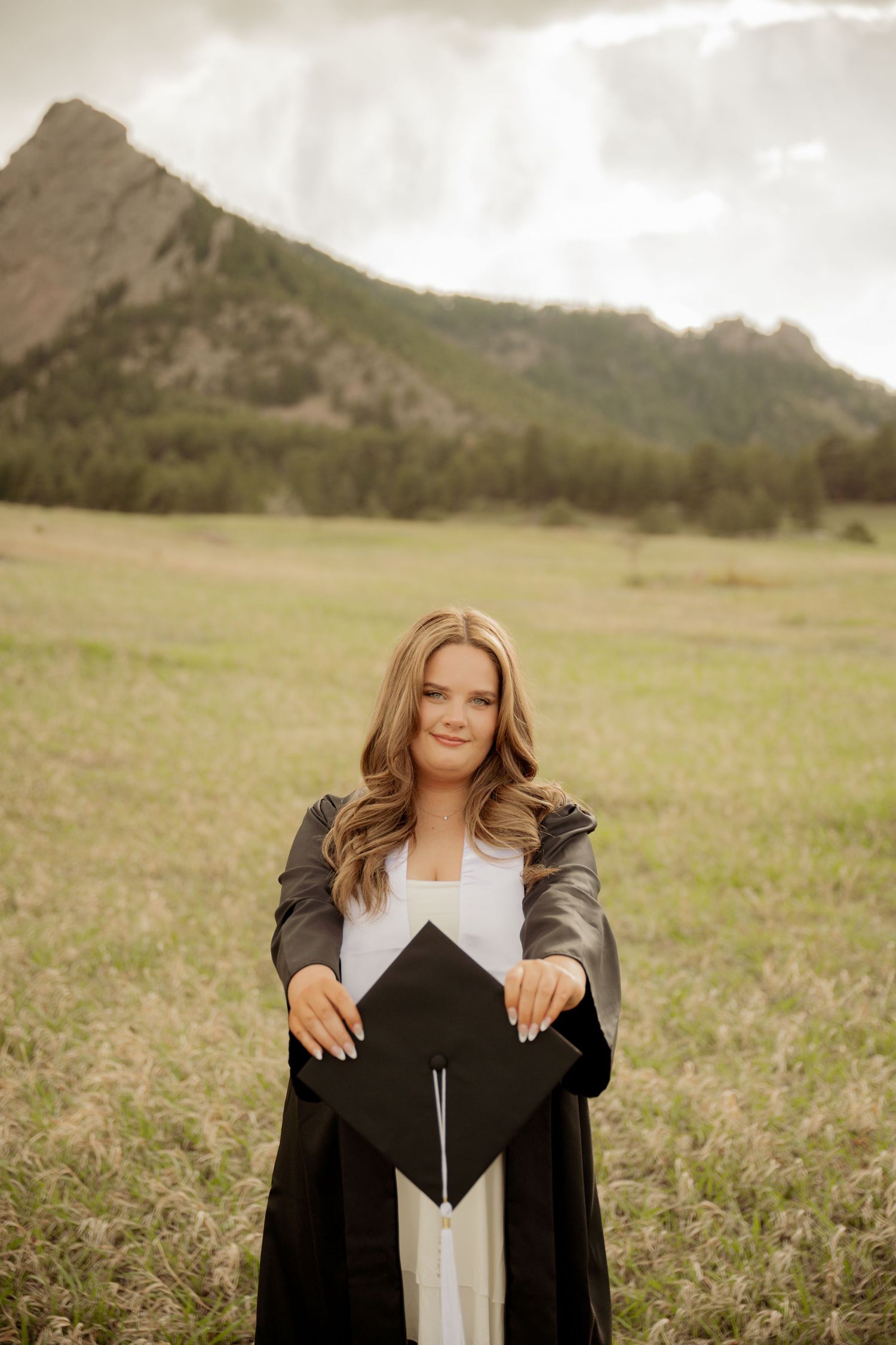 A graduation portrait in a meadow with mountains in the background shows someone holding a black cap in golden light.