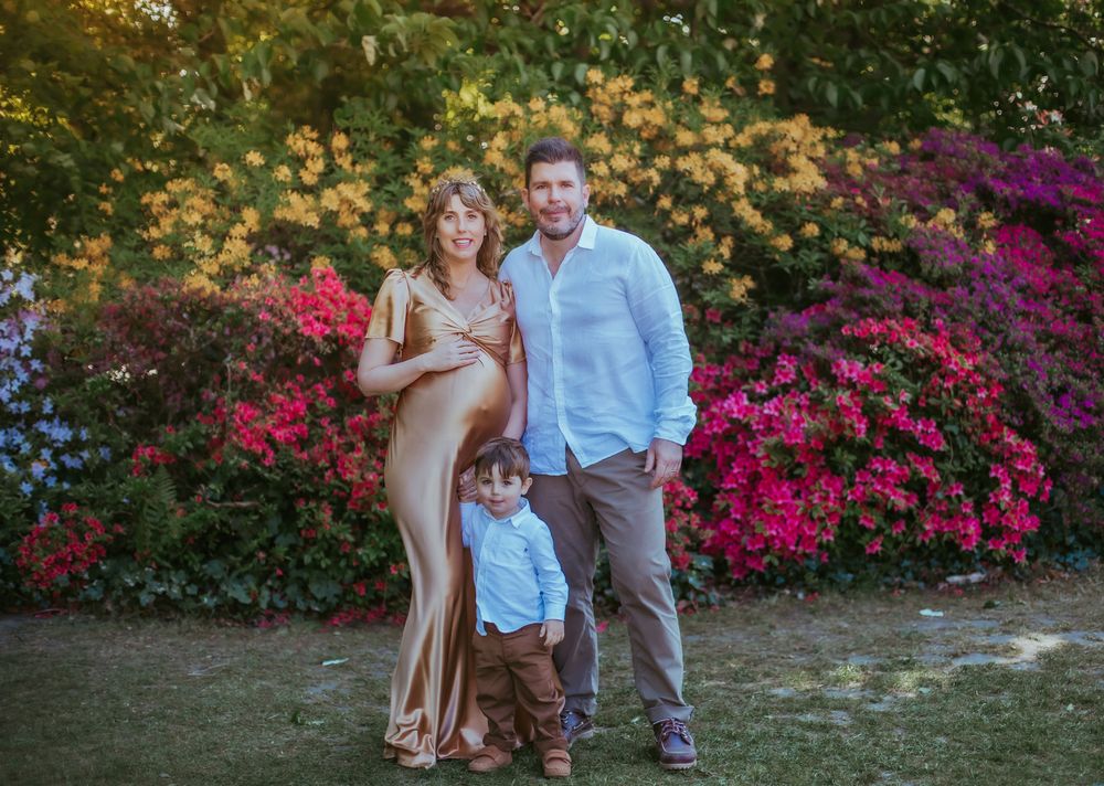 Family stands together in front of vibrant pink and yellow flowers in a garden setting at sunset. During a maternity session in London
