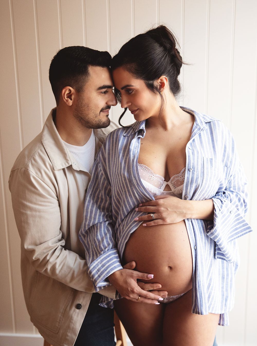 Intimate maternity photo of couple sharing tender moment with hands on pregnant belly in striped shirt and beige jacket.