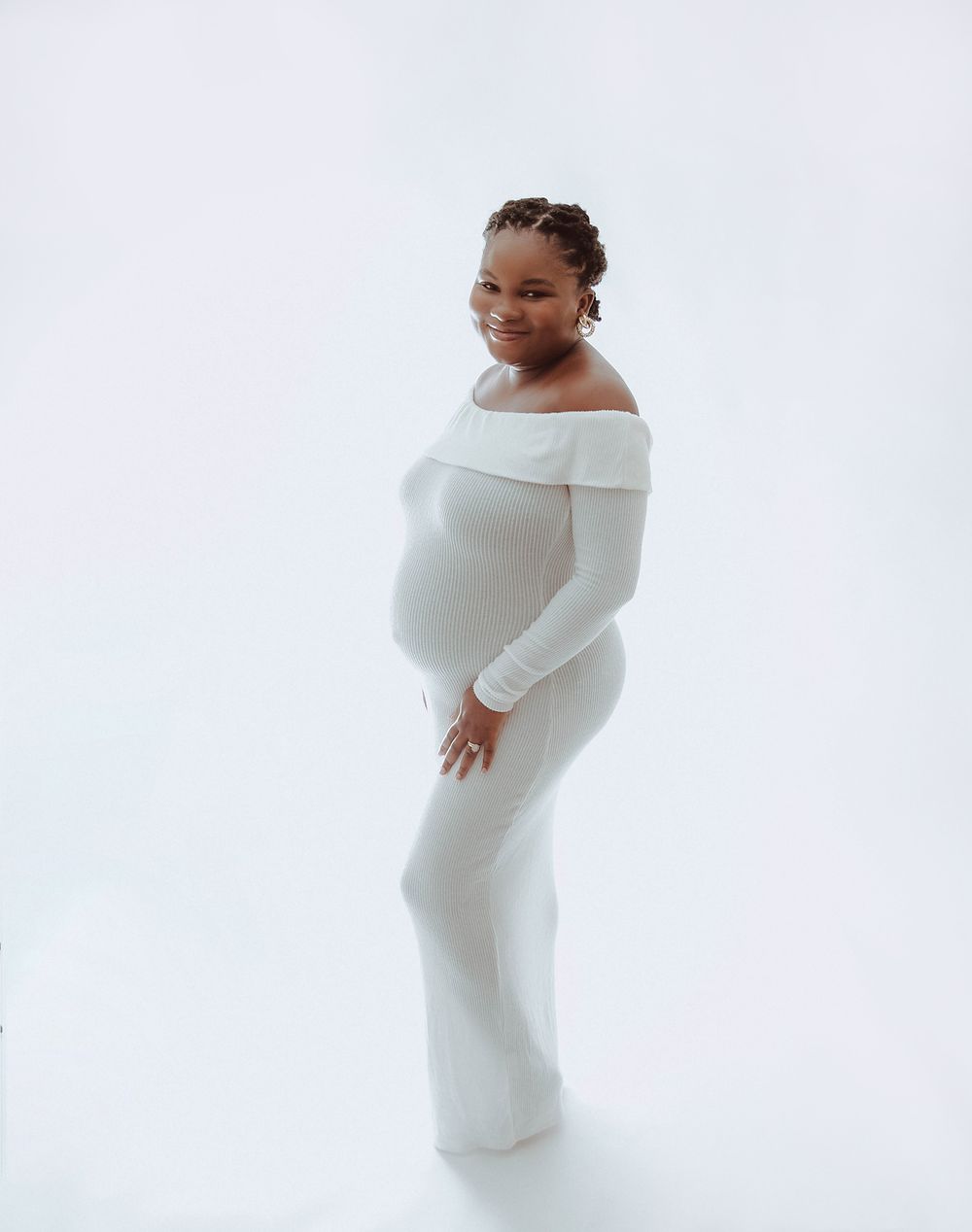 Expectant person poses in elegant white form-fitting off-shoulder maternity gown against bright white background. During a maternity session