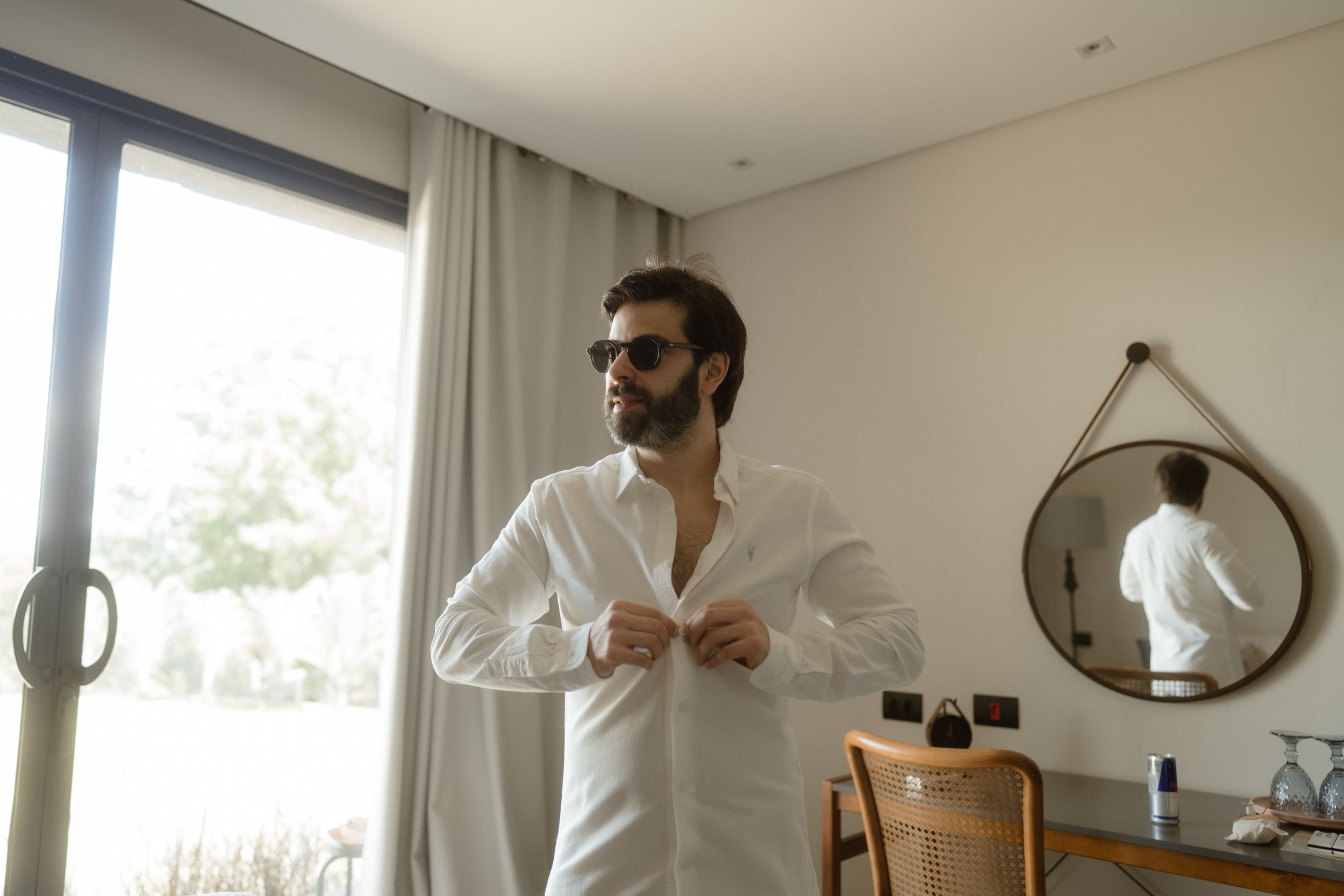 A person getting dressed in a white shirt near a circular mirror in a modern bedroom.