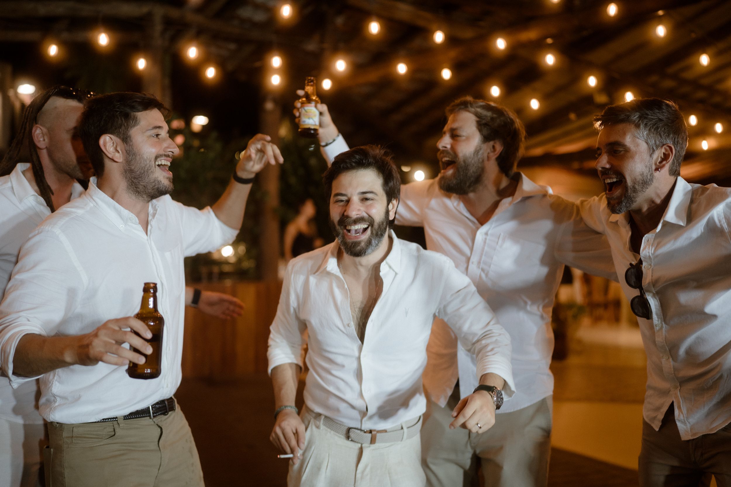 Wedding celebration guests in white shirts enjoy dancing under festive string lights during evening reception.