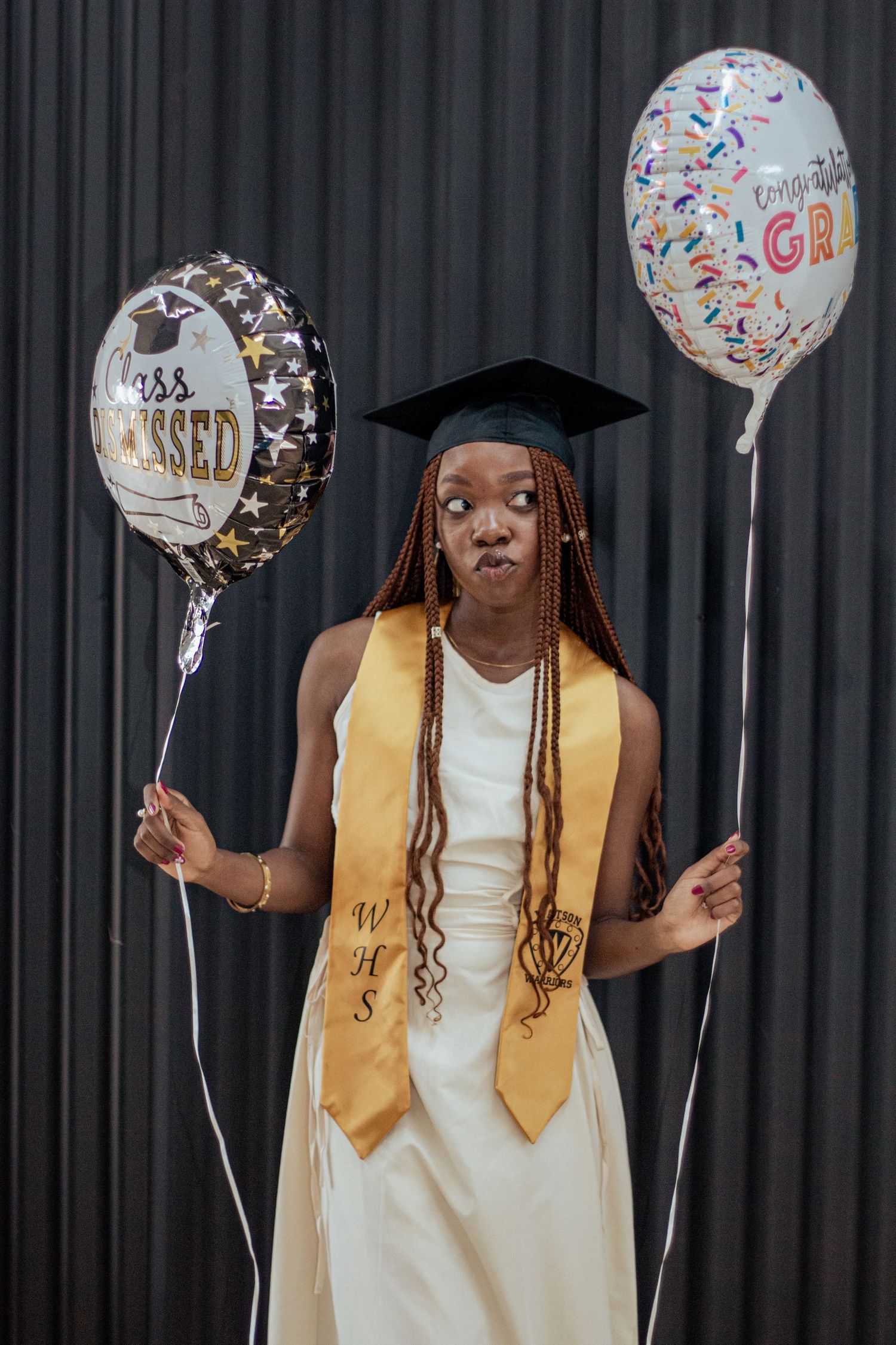 Graduate in gold stole and cap holds two celebratory balloons against grey background at commencement ceremony.