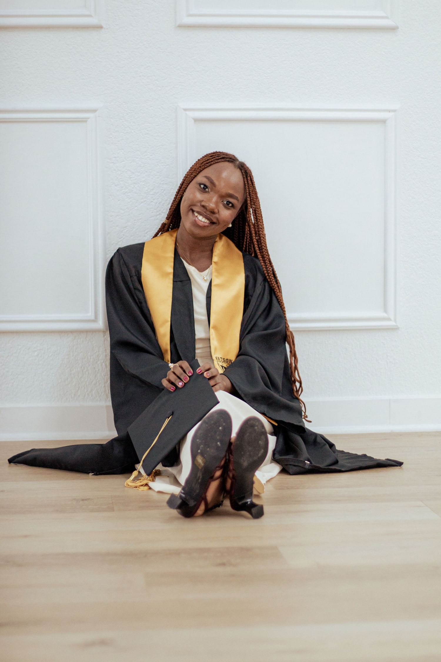 Graduate wearing black and gold academic regalia sits on hardwood floor with tilted head against white paneled wall.