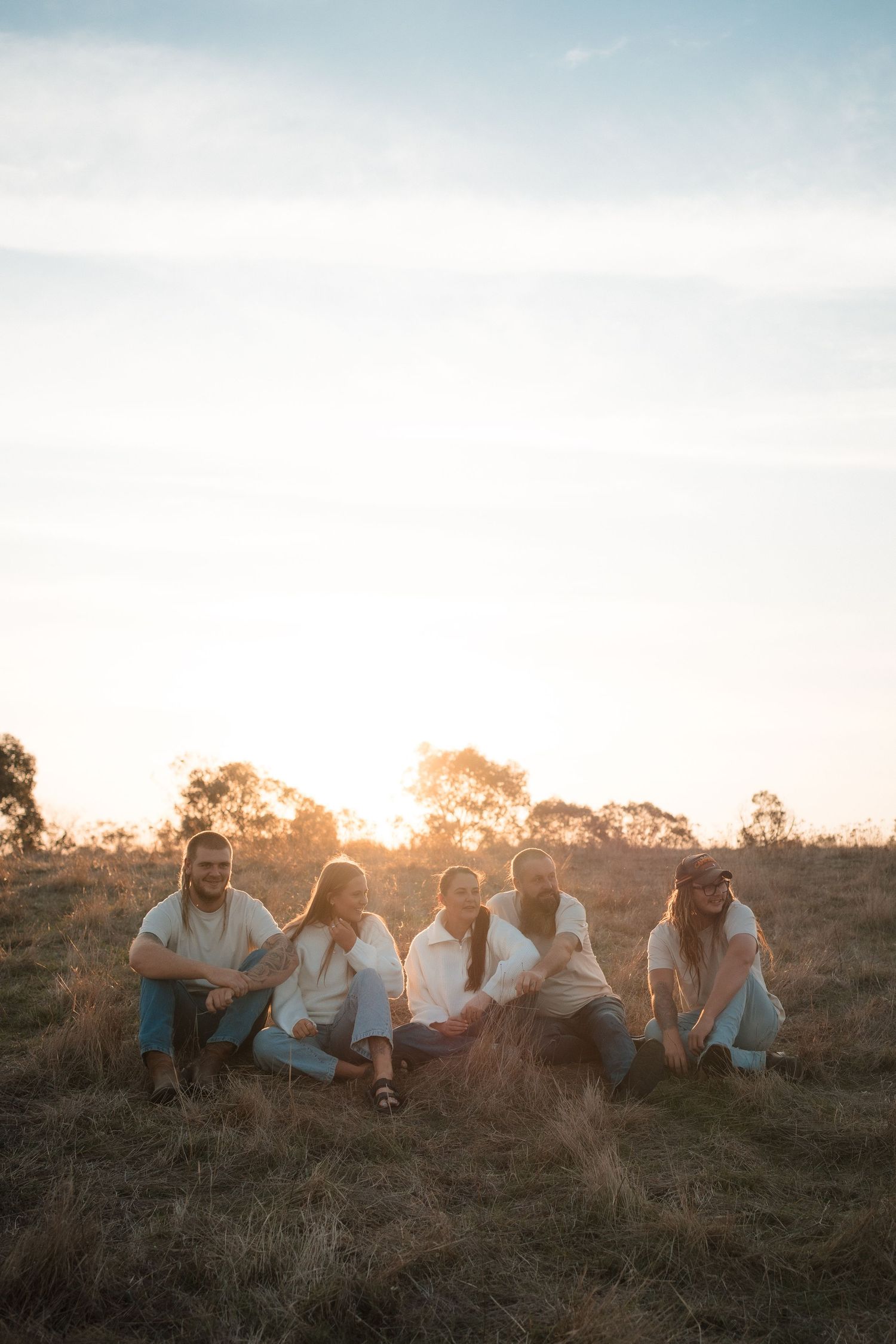 Group of people sitting together on grassy hill at sunset in casual white clothing enjoying peaceful moment outdoors.
