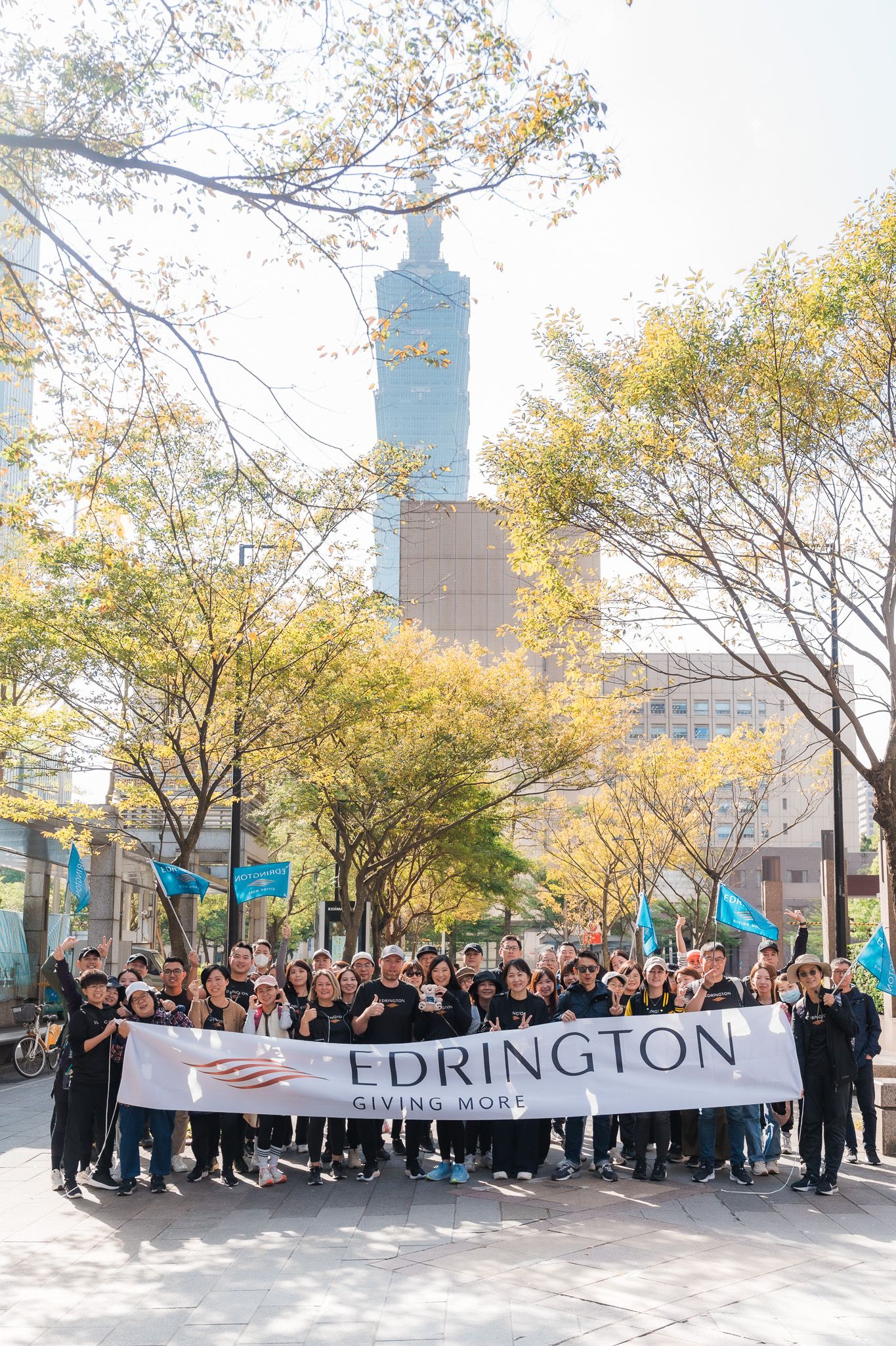 A large group of people holding an Edrington banner gather in front of Taipei 101 surrounded by autumn trees.