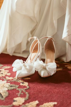 Elegant white bridal shoes with delicate bows sit on a red and gold patterned carpet beside flowing white fabric.