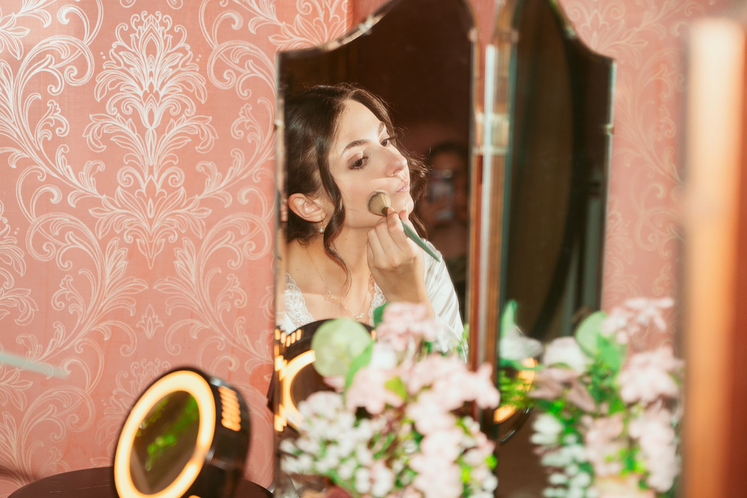 Woman in bridal robe touching up lipstick at vintage-styled vanity with pink floral wallpaper and flowers.
