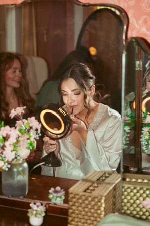 A bridal preparation scene with someone applying makeup using an illuminated mirror surrounded by floral arrangements.
