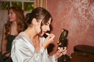 A person in a silk robe applies lipstick while looking in a compact mirror during a preparation event.