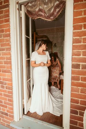 A person in a white mermaid-style wedding dress stands in a doorway of a rustic brick building.