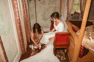 Two people help arrange a white wedding dress in an ornate room with vintage decor and wooden banister.
