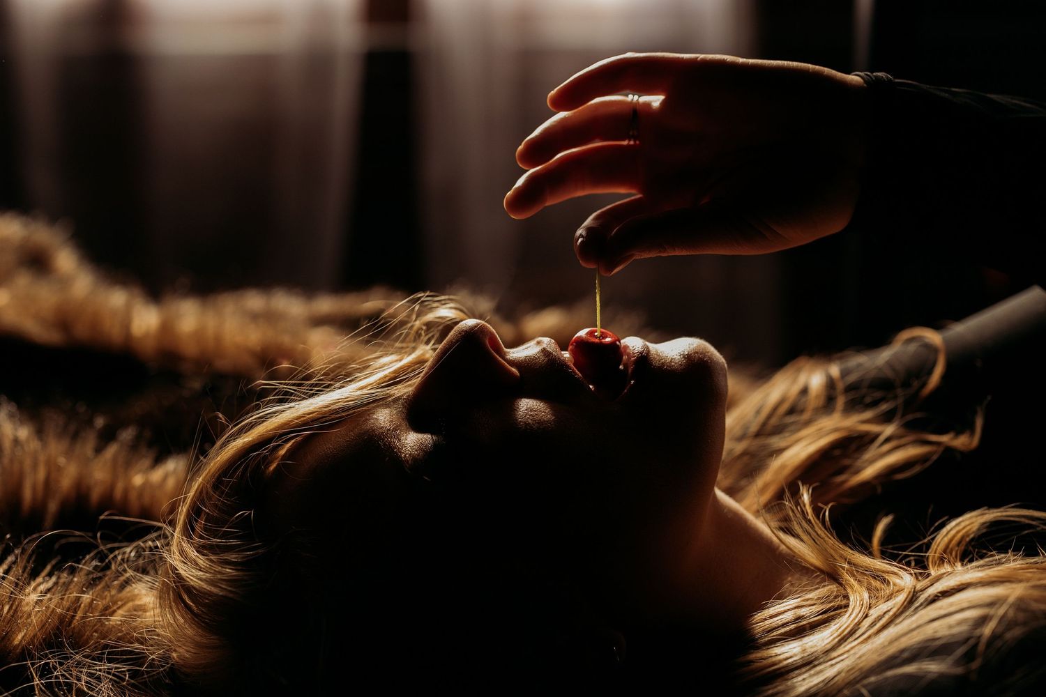 Dark silhouette of a person in a boudoir pose kneeling on carpet near window with brick wall and white curtains.