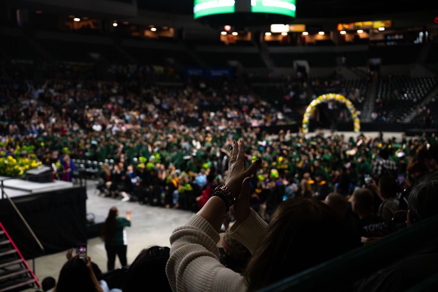 Crowd at a graduation ceremony in a large arena, with green and gold decorations and a stage visible in the background.