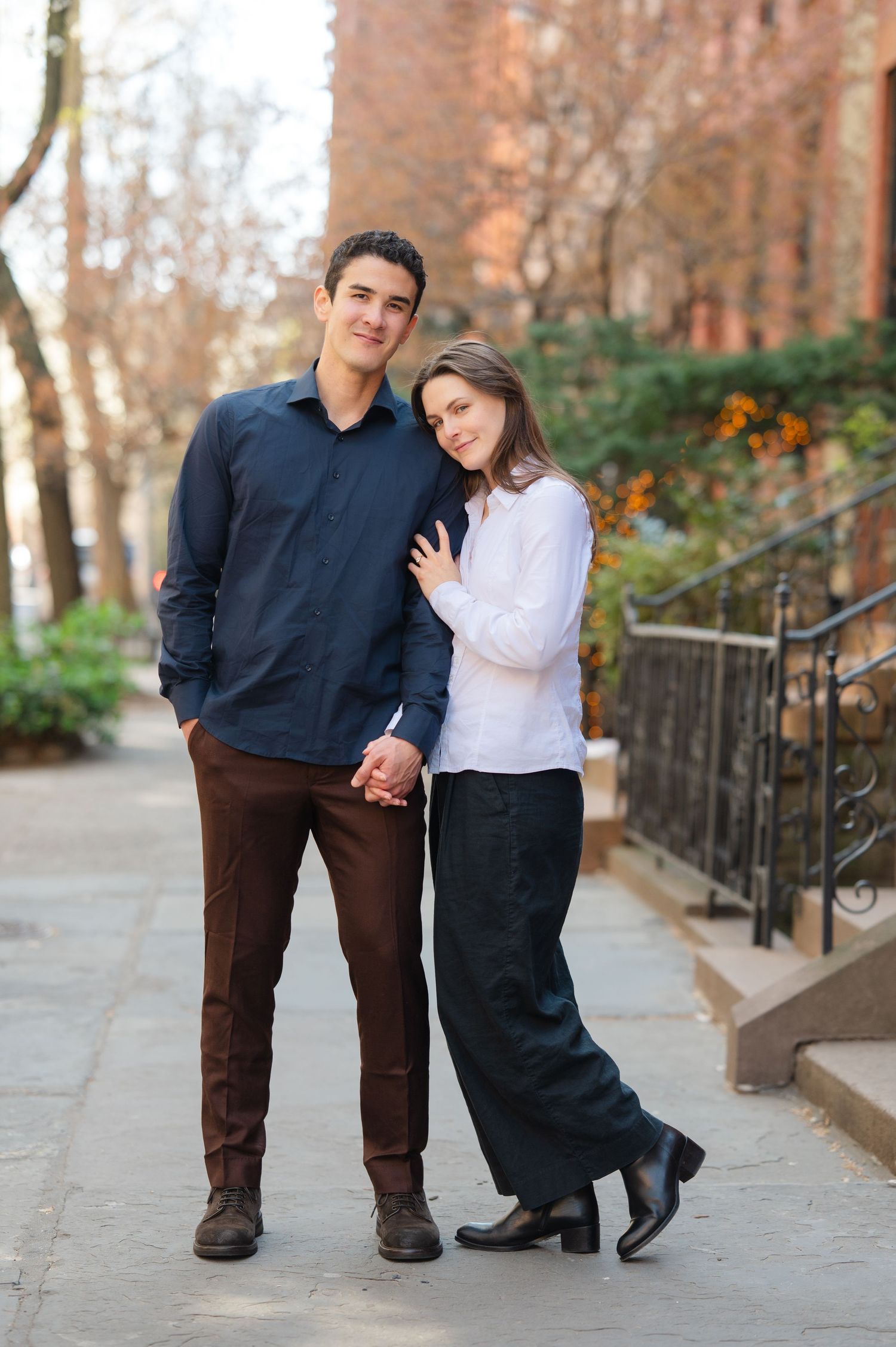 A couple poses together on a tree-lined urban sidewalk, the woman leaning on the man's shoulder during a proposal session in NYC