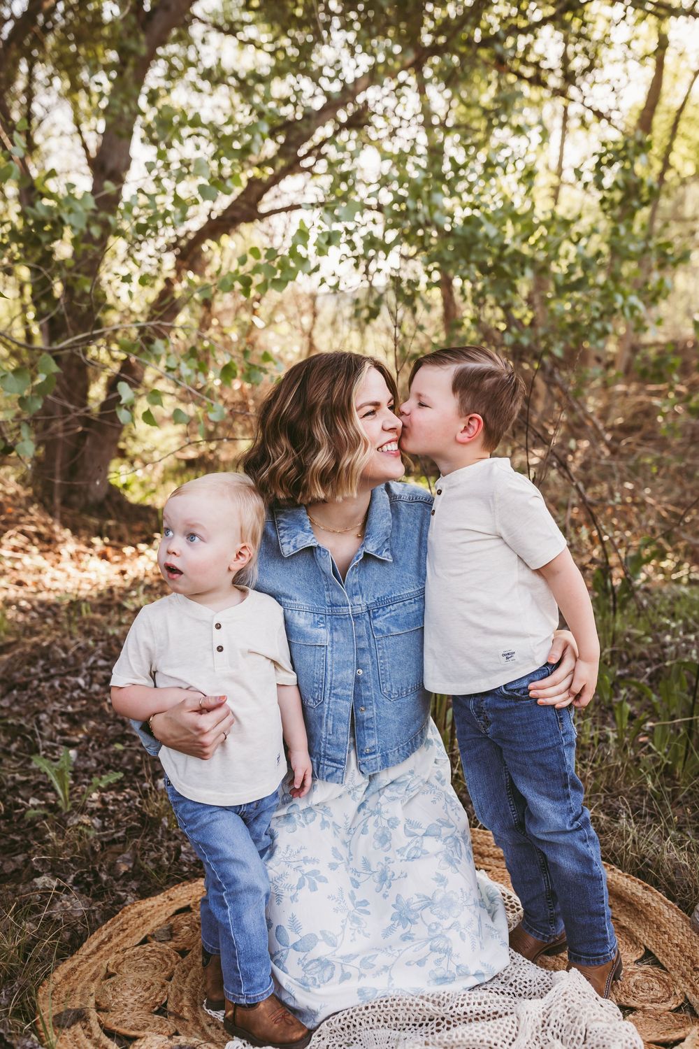 A mother sits outdoors with two young boys, one kissing her cheek, surrounded by lush green trees for mommy and me photos in Beaumont, Ca.