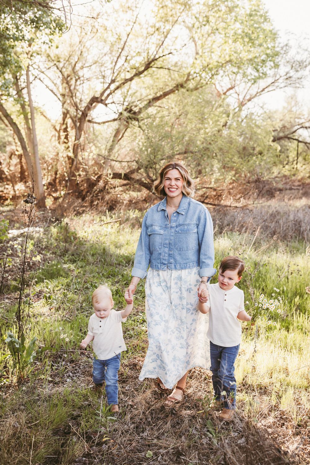 A smiling woman in a denim jacket and floral dress walks outdoors holding hands with two young boys during a spring session.