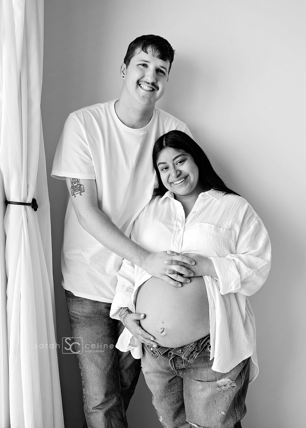 Black and white maternity photo of a smiling couple, woman cradling baby bump, both wearing white shirts and jeans.