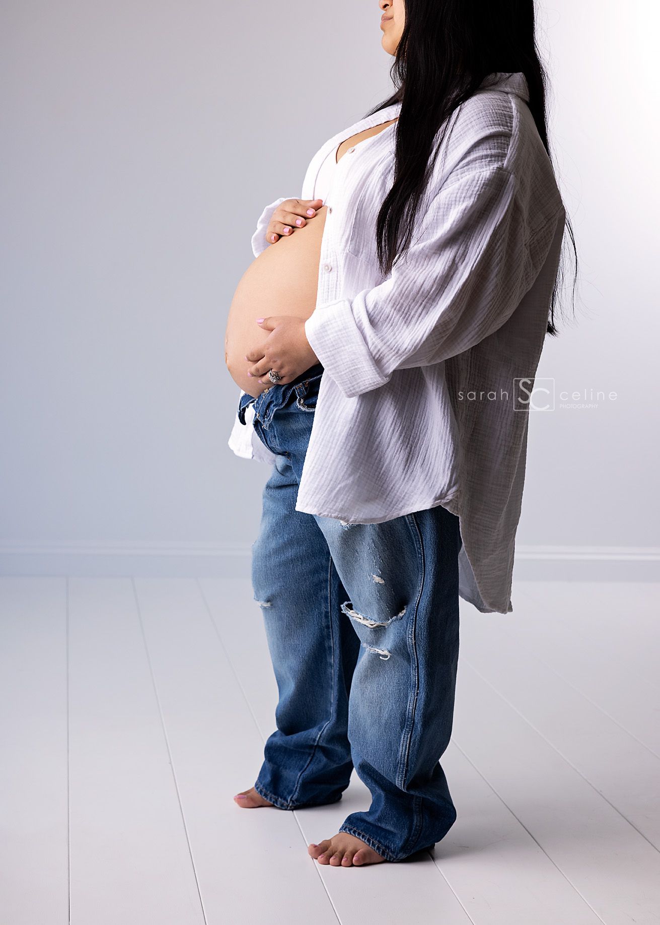 Pregnant woman in open white shirt and ripped jeans showing baby bump in a bright studio maternity photo.
