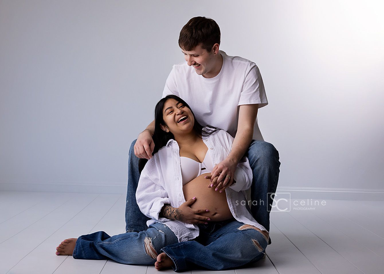 Couple sharing a joyful maternity photo session on studio floor, woman laughing as partner touches her baby bump.