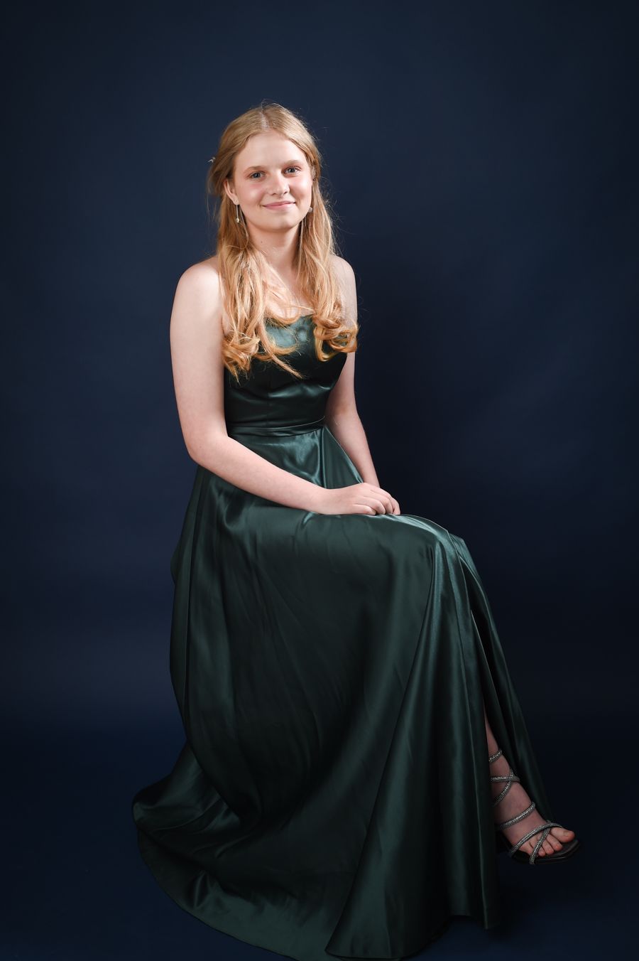 Young woman in an elegant dark green gown, seated against a dark blue backdrop. She has long blonde hair and a gentle smile, conveying a ser