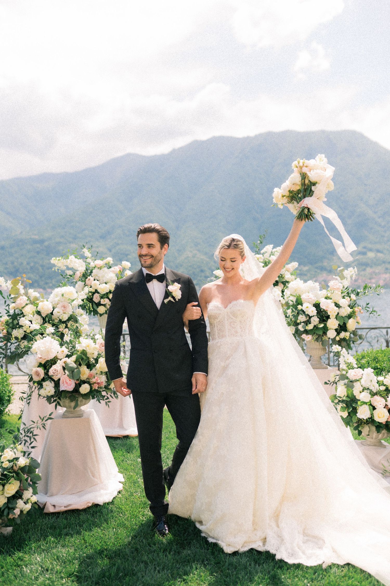 Bride and groom sharing a romantic kiss under a flowing white veil, with a lush white floral bouquet in the foreground.