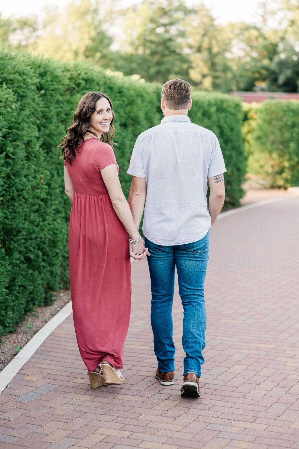 Couple holding hands walking on brick path, woman in red maxi dress looks back, man in white shirt and jeans.