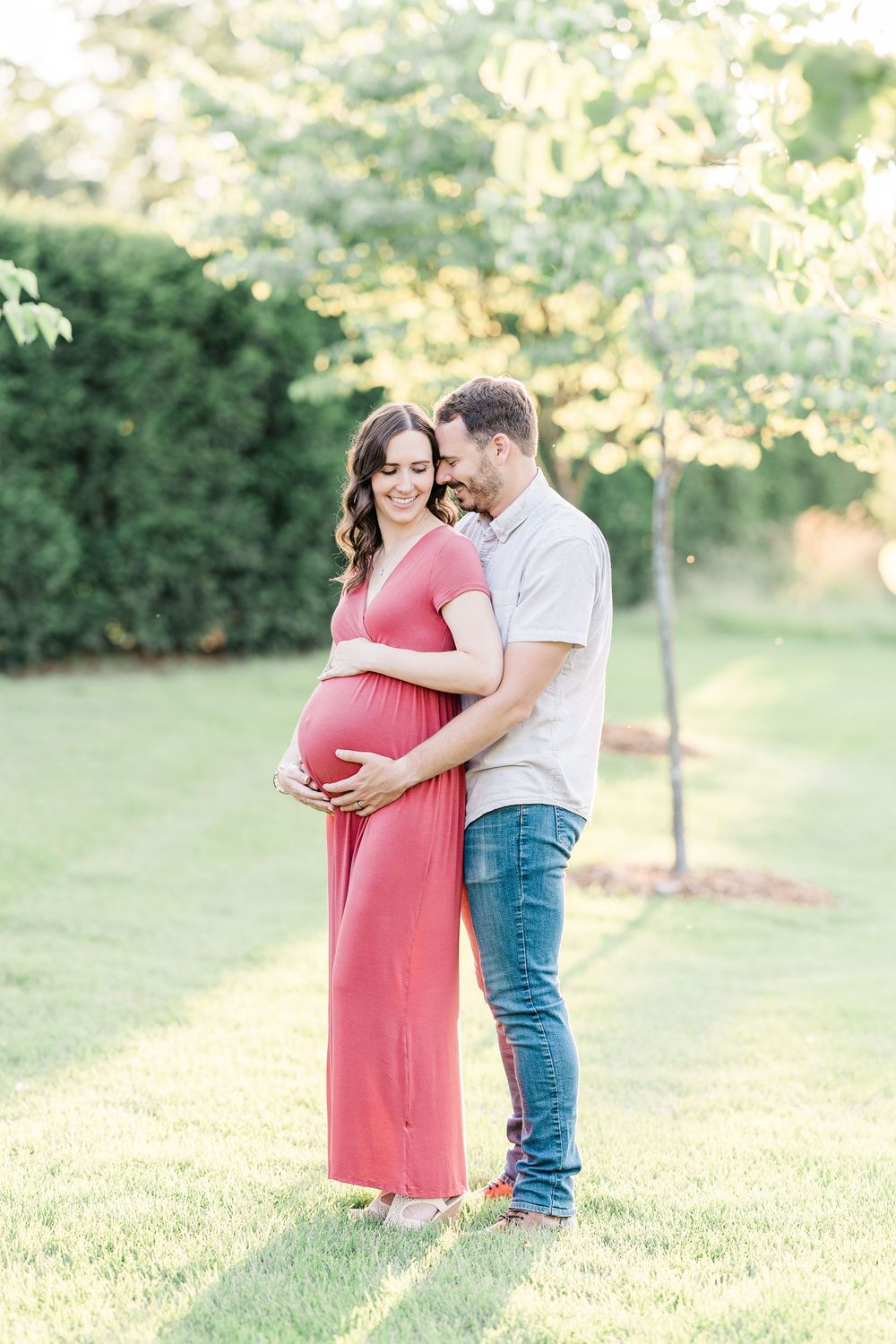 Pregnant woman in red maxi dress embraced by partner in sunny green park during maternity photo session.