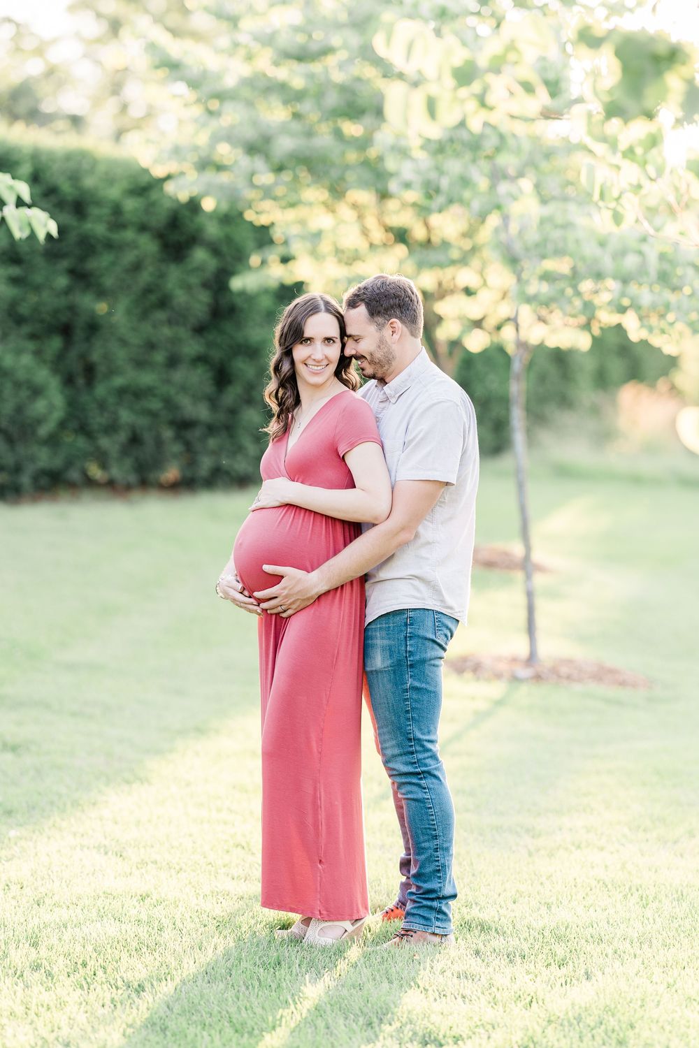 Smiling pregnant woman in coral dress held by partner outdoors in lush garden maternity portrait.