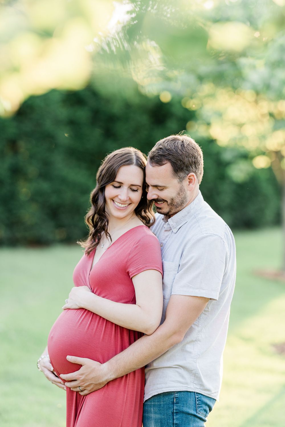 Smiling pregnant woman in red dress embraced by man outdoors in sunny green park during maternity photo session.