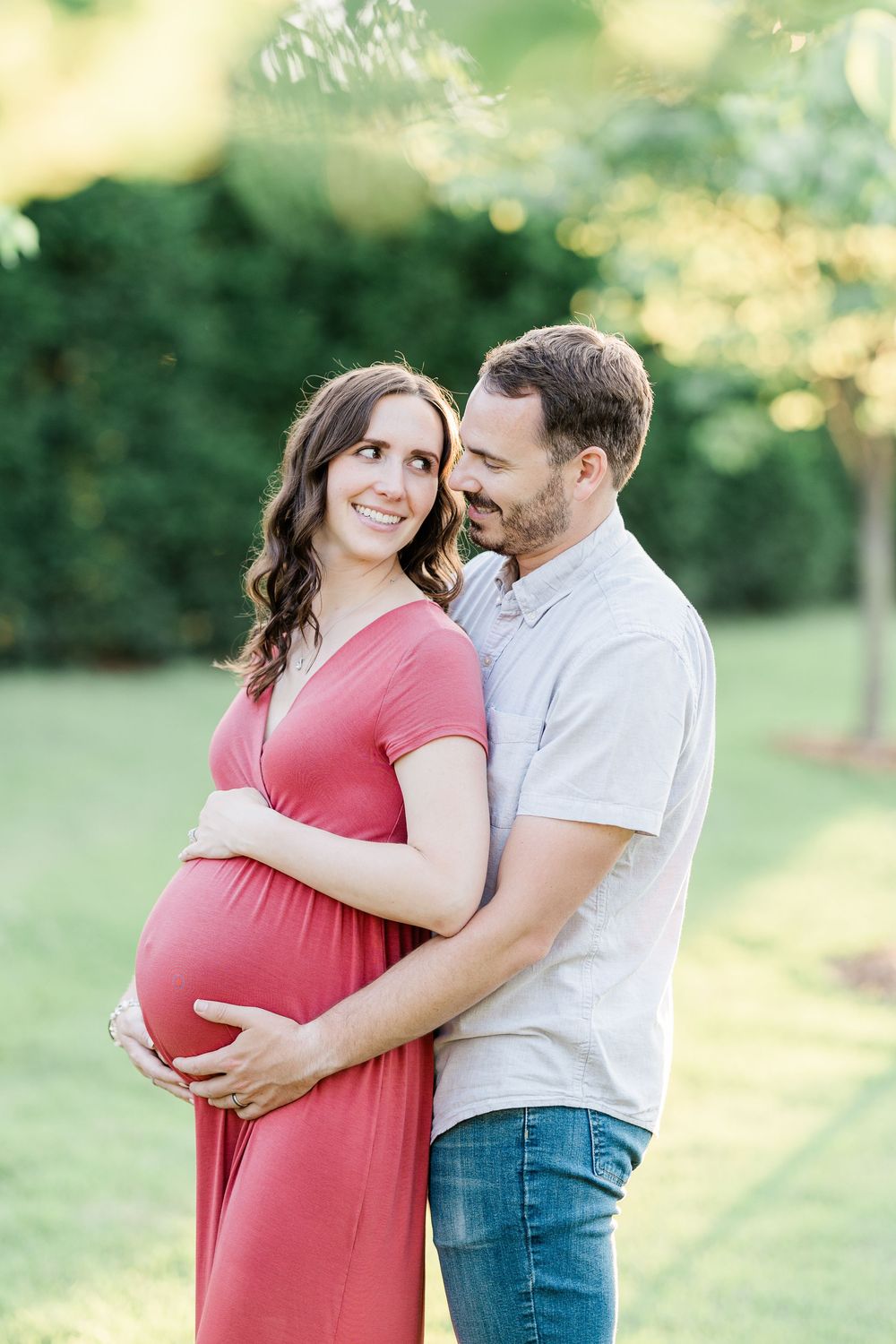 Happy expectant couple embracing in park, woman in pink dress cradling baby bump during maternity shoot.