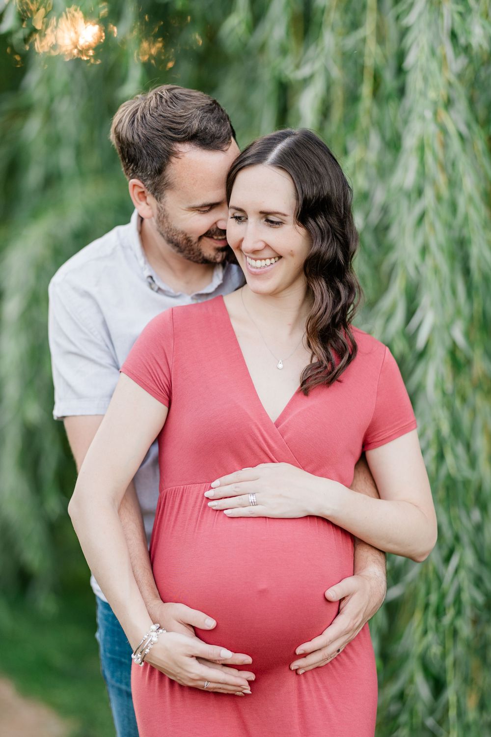 Smiling pregnant woman in red dress embraced by partner under weeping willow tree.