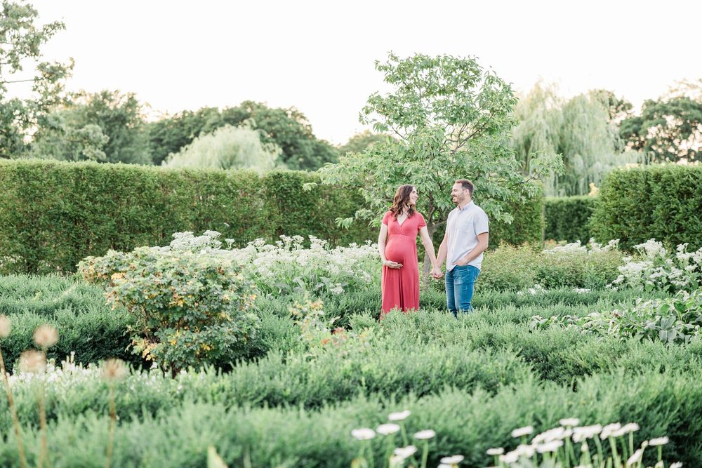 Couple holding hands in lush garden, woman in red maxi dress and man in white shirt surrounded by greenery.