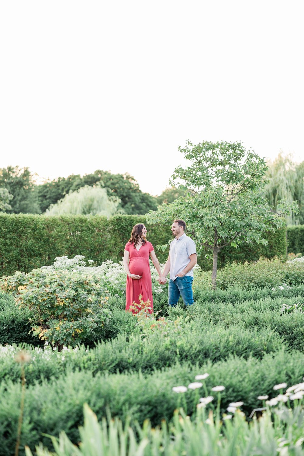 Pregnant couple holding hands in a lush garden, woman in red dress, man in gray shirt and jeans.