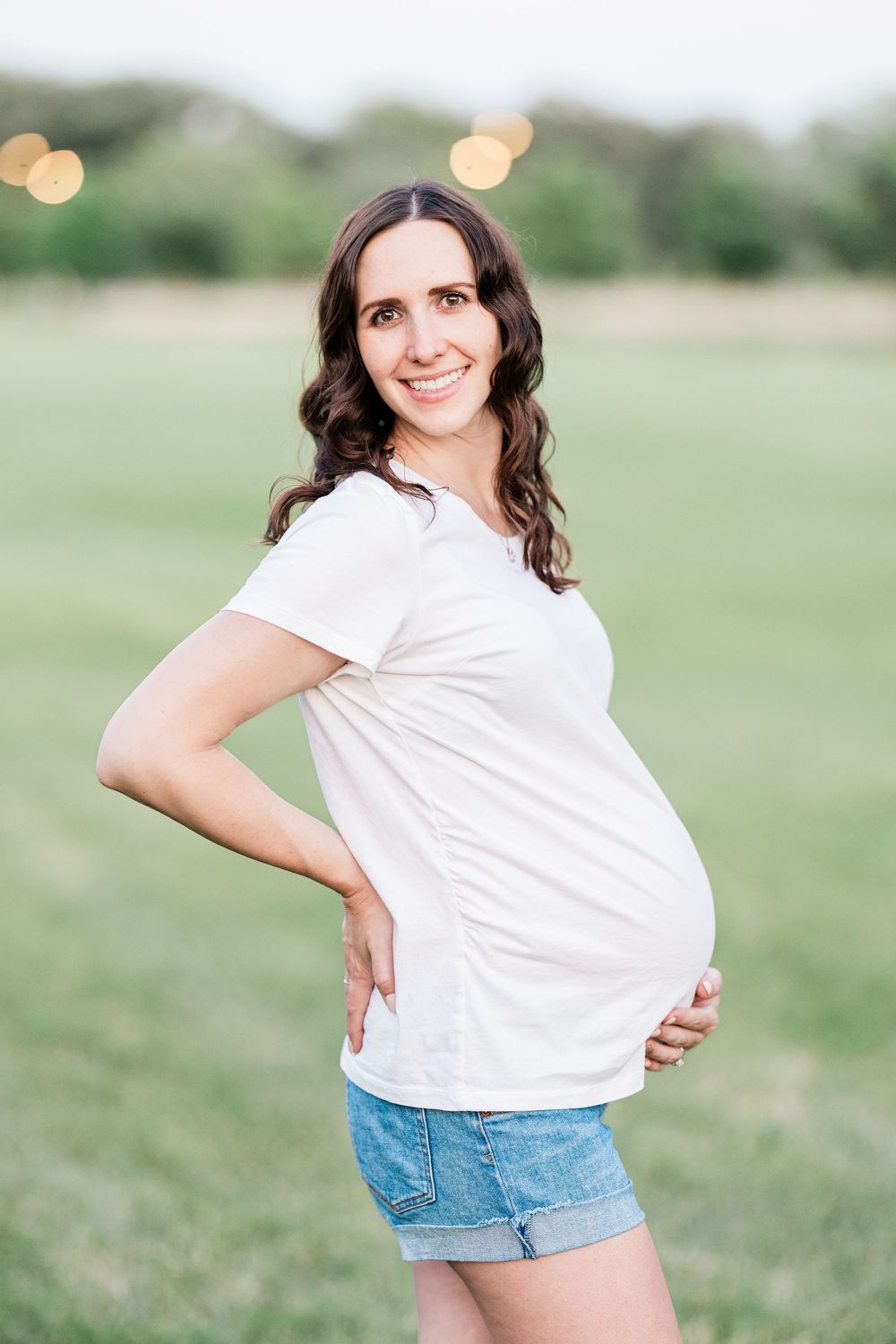 Happy pregnant woman in white tee and denim shorts cradling baby bump in sunny outdoor field.