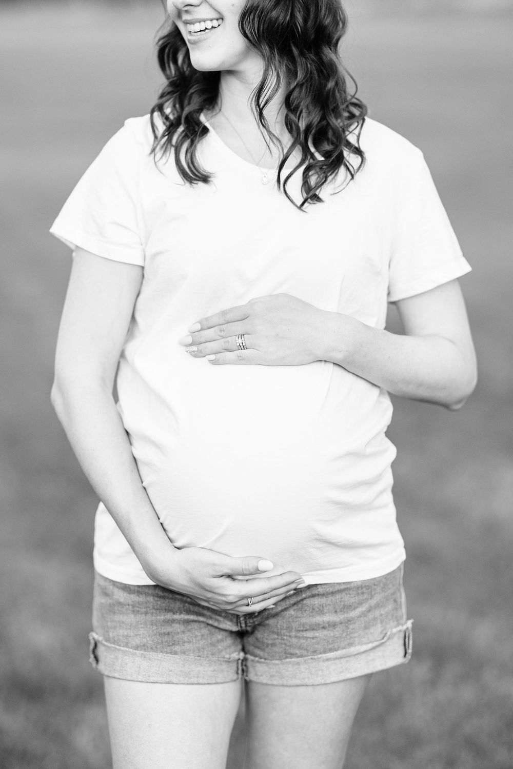Black and white photo of a smiling pregnant woman in a white t-shirt and denim shorts cradling her baby bump outdoors.