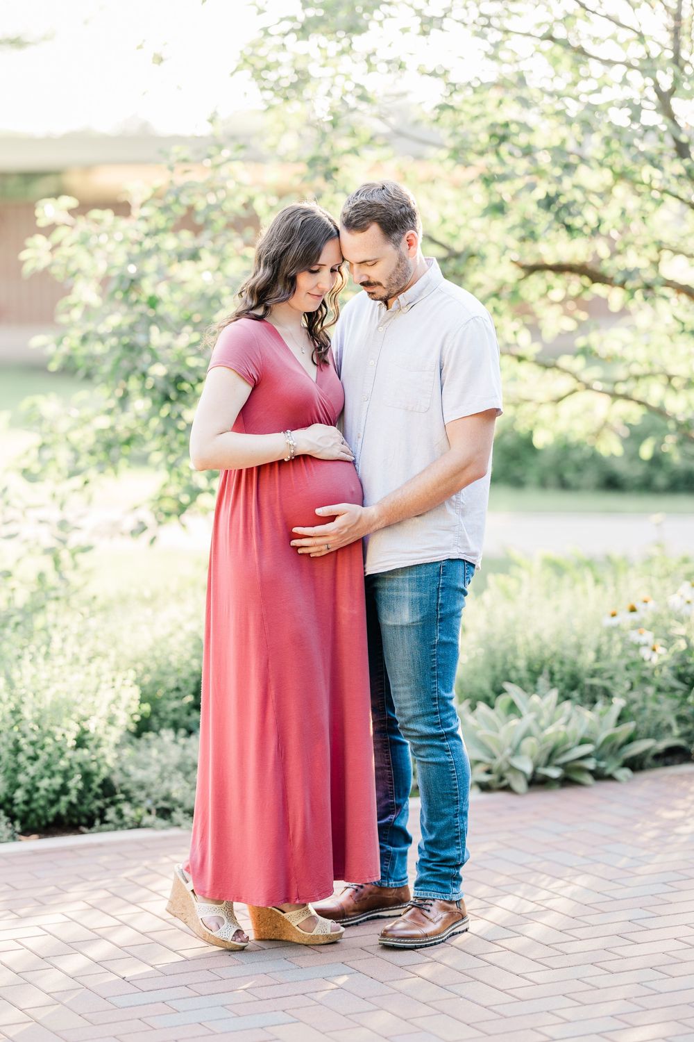 A couple tenderly gazes at the pregnant woman's belly during an outdoor maternity photo session in a garden setting.