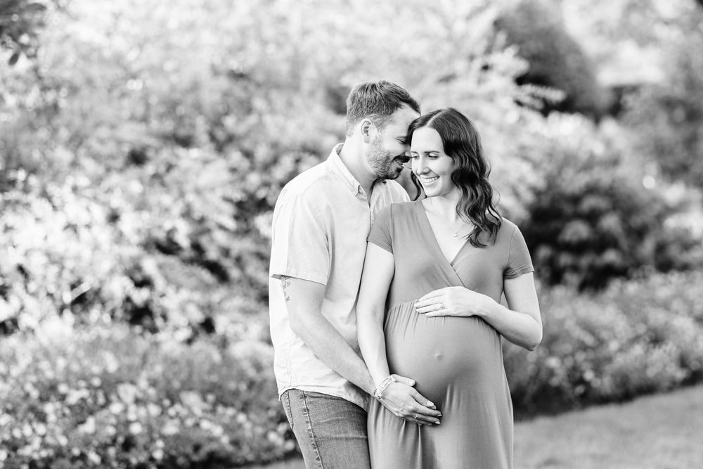 Black and white maternity photo of a couple outdoors, woman in flowing dress cradling baby bump while partner gazes lovingly.