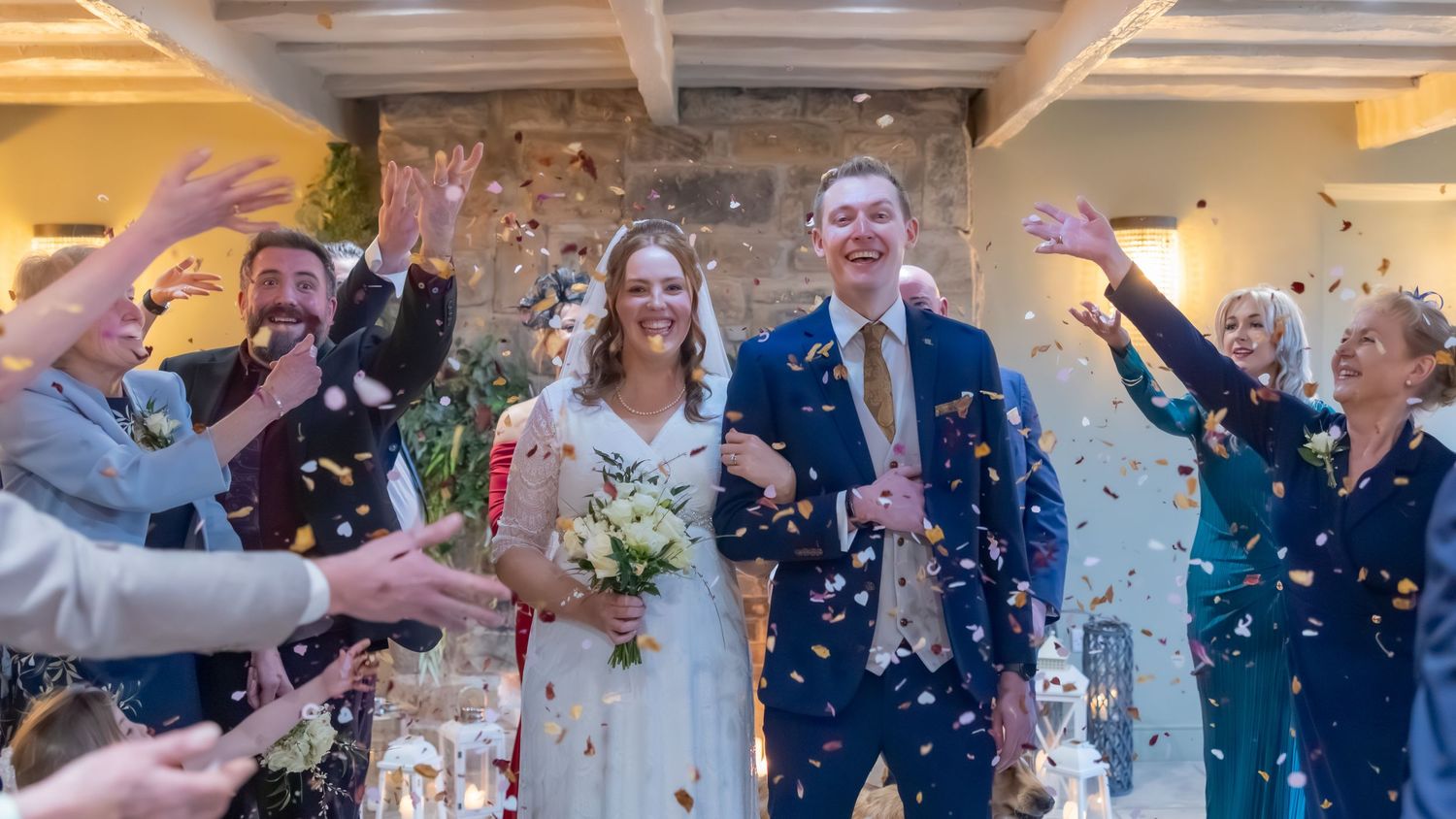 Bride and groom walk through confetti shower surrounded by cheering guests at elegant indoor wedding ceremony. Casual Captures Wedding