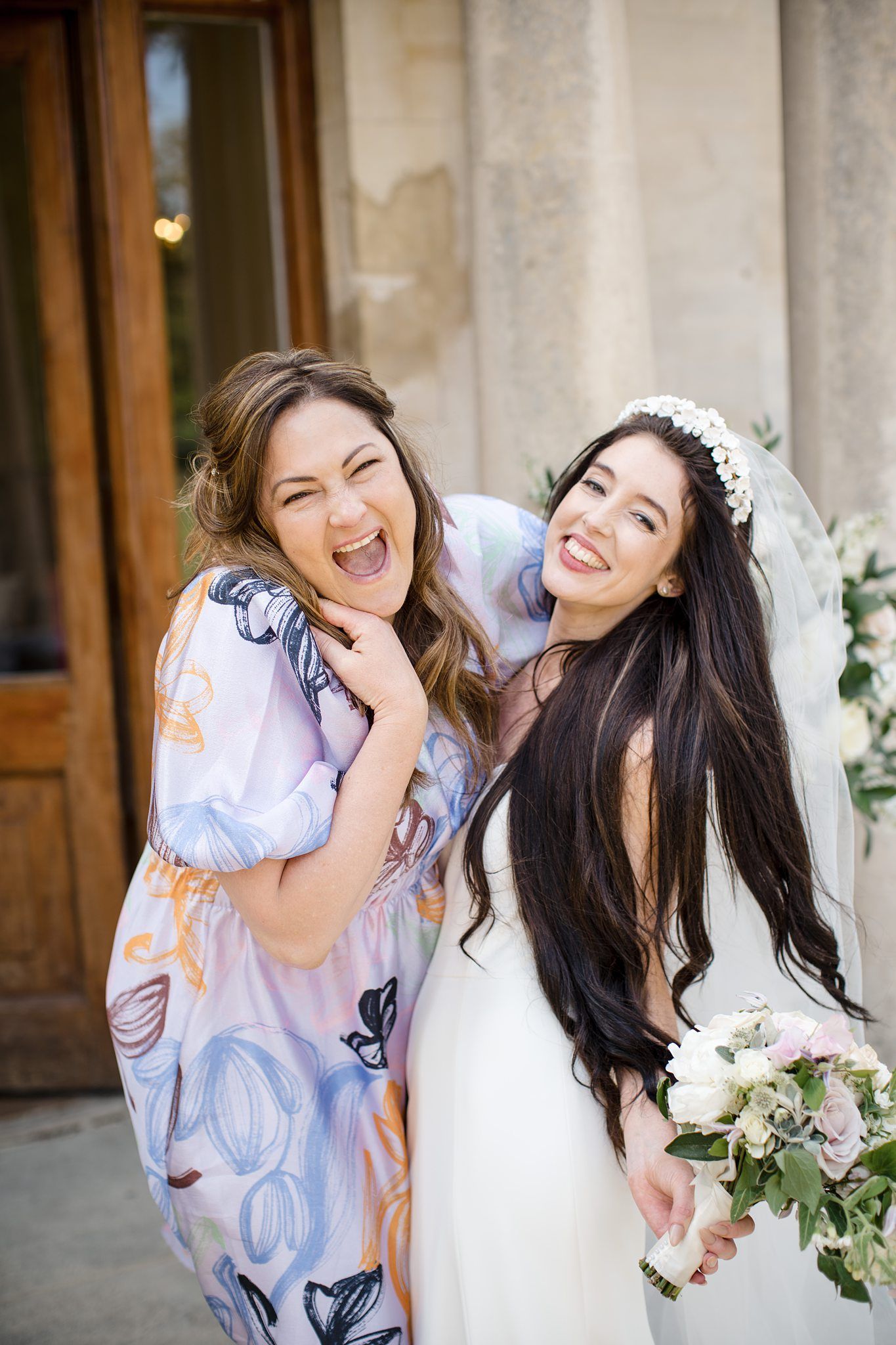 Bride in white gown and veil holds bouquet while laughing with friend in floral dress outside church.