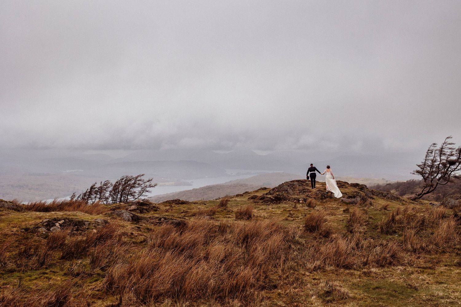 A bride and groom stand on a rugged hilltop amid golden grass under a dramatic overcast sky in a remote landscape.